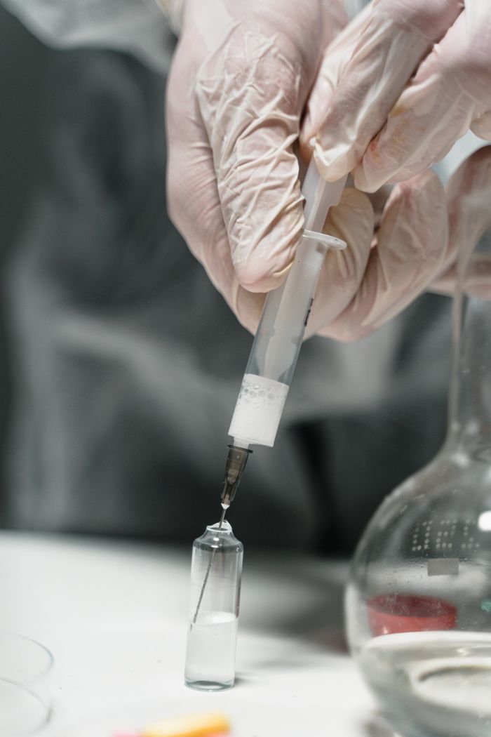services-02 Close-up of hands in gloves using a syringe to extract liquid from a vial in a lab setting.